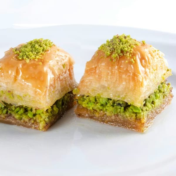 Two pieces of golden-brown baklava, topped with crushed green pistachios, sit on a white plate.