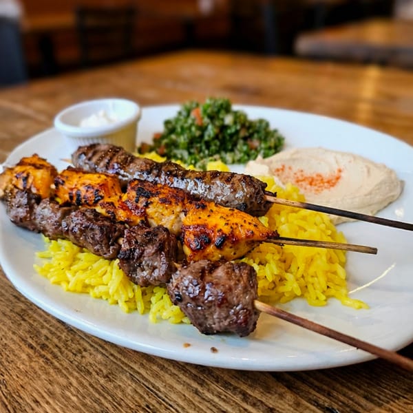Grilled chicken and beef skewers with yellow rice, hummus, and tabbouleh salad on a white plate.