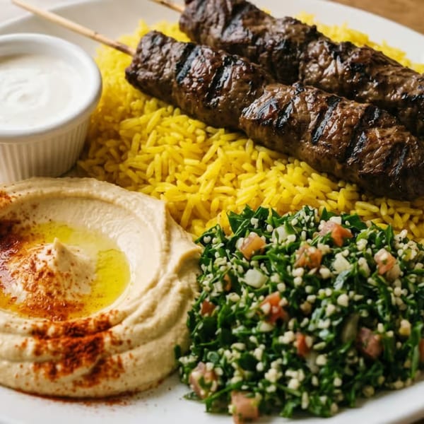 Two grilled meat skewers on yellow rice, with hummus, tabbouleh salad, and a white sauce.
